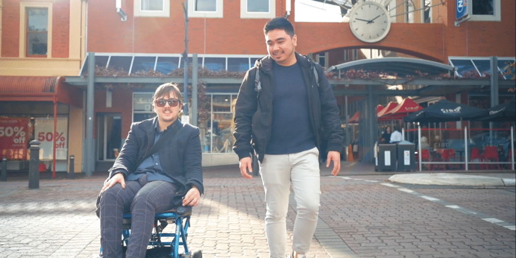 Jason in a wheelchair and another man walking beside him smile as they move through a sunny urban area with shops and a large clock in the background.